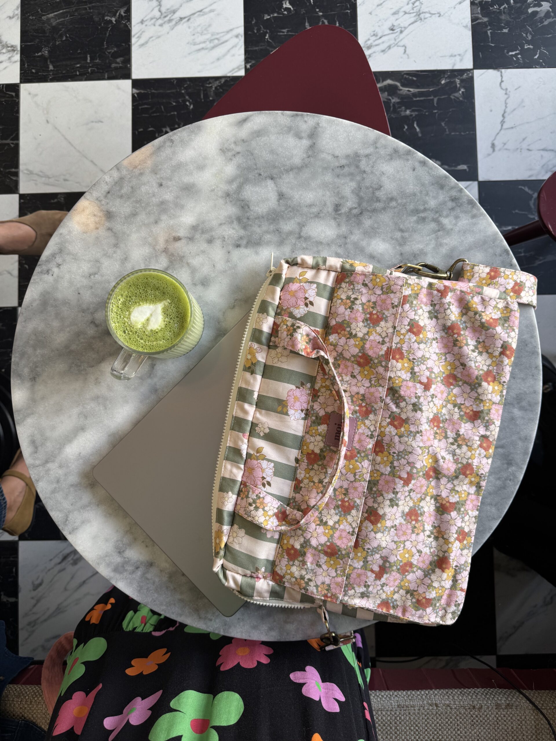 Okrągły marmurowy stolik na tle czarno-białej szachownicy, na stoliku laptop podłożony pod kolorową, kwiatową torbą i filiżanka zielonej herbaty latte z pianką w kształcie serca, widoczne fragmenty kolorowej sukienki użytkownika i obuwia.