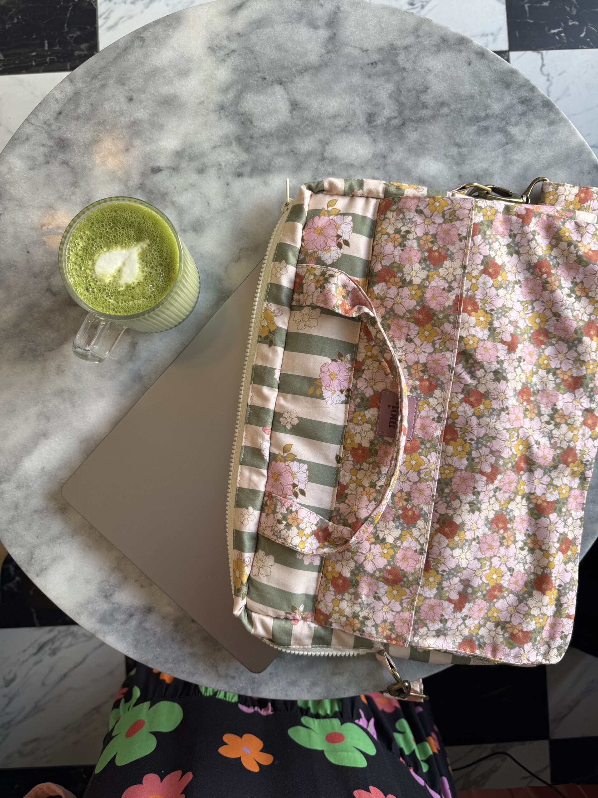 Stolik z marmurowym blatem, na którym leży zamknięty różowy laptop częściowo przykryty kwiecistą torbą na laptopa w pastelowych kolorach; obok stoi szklanka z zielonym napojem z pianką w kształcie liścia; u dołu zdjęcia widoczna część spódnicy z kolorowymi nadrukami kwiatowymi; tło to czarno-biała podłoga w kratę.