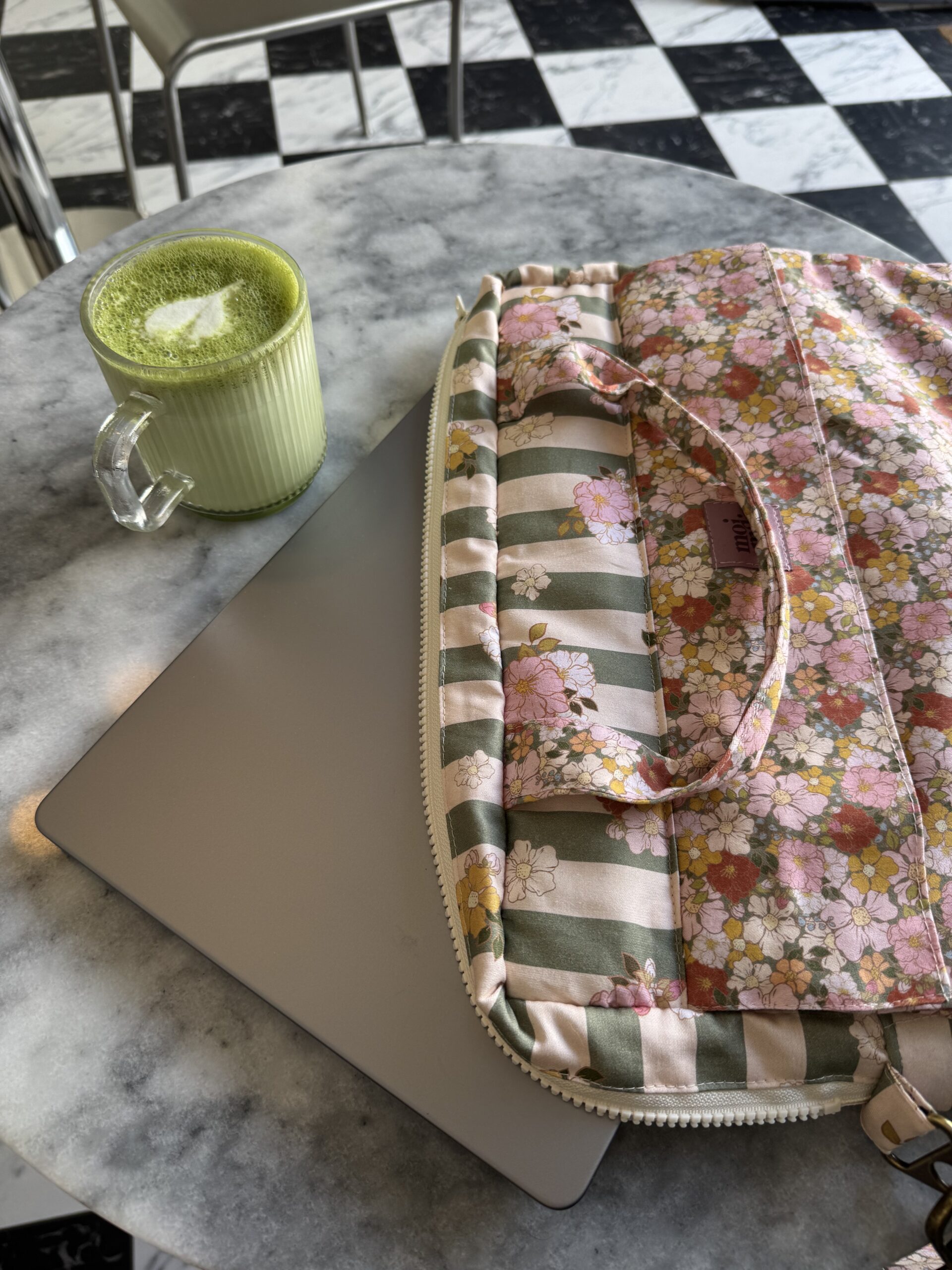 Szary laptop częściowo w różowo-zielonym kwiatowym i paskowanym pokrowcu na marmurowym stole obok szklanki z zielonym napojem z pianką i rysunkiem liścia na wierzchu, w tle krzesło i podłoga w czarno-białą kratkę.