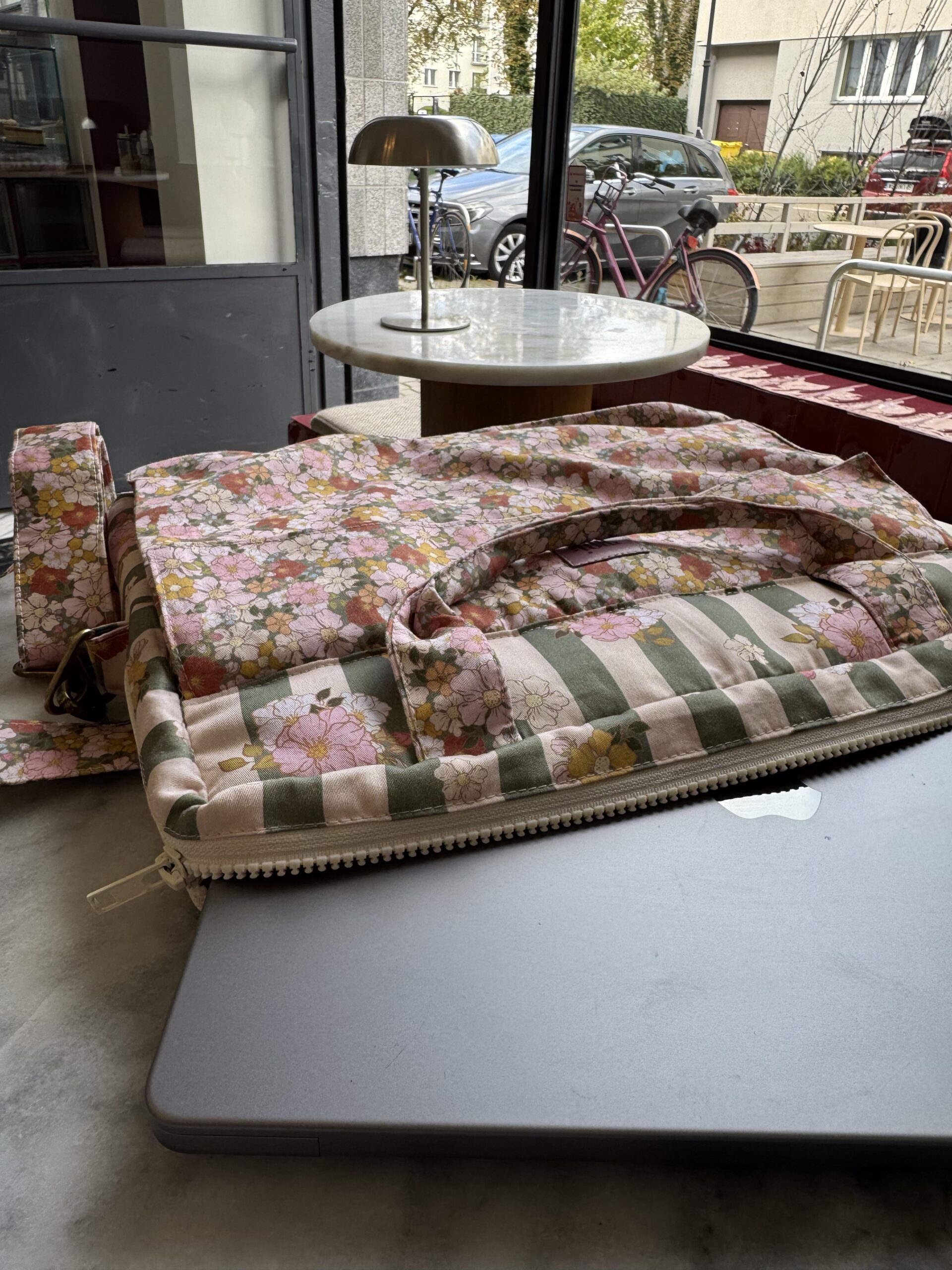 Szara zamknięta laptopa częściowo schowana w kwiatowej torbie z dużym paskiem, na stole w kawiarni, w tle okno z widokiem na rower i samochód oraz stół z lampą stołową