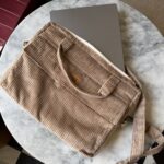 Beżowa torba z materiału sztruksowego częściowo zasłaniająca szary laptop na okrągłym marmurowym stole, w tle czerwona tapicerka i czarno-biała podłoga w kafle.