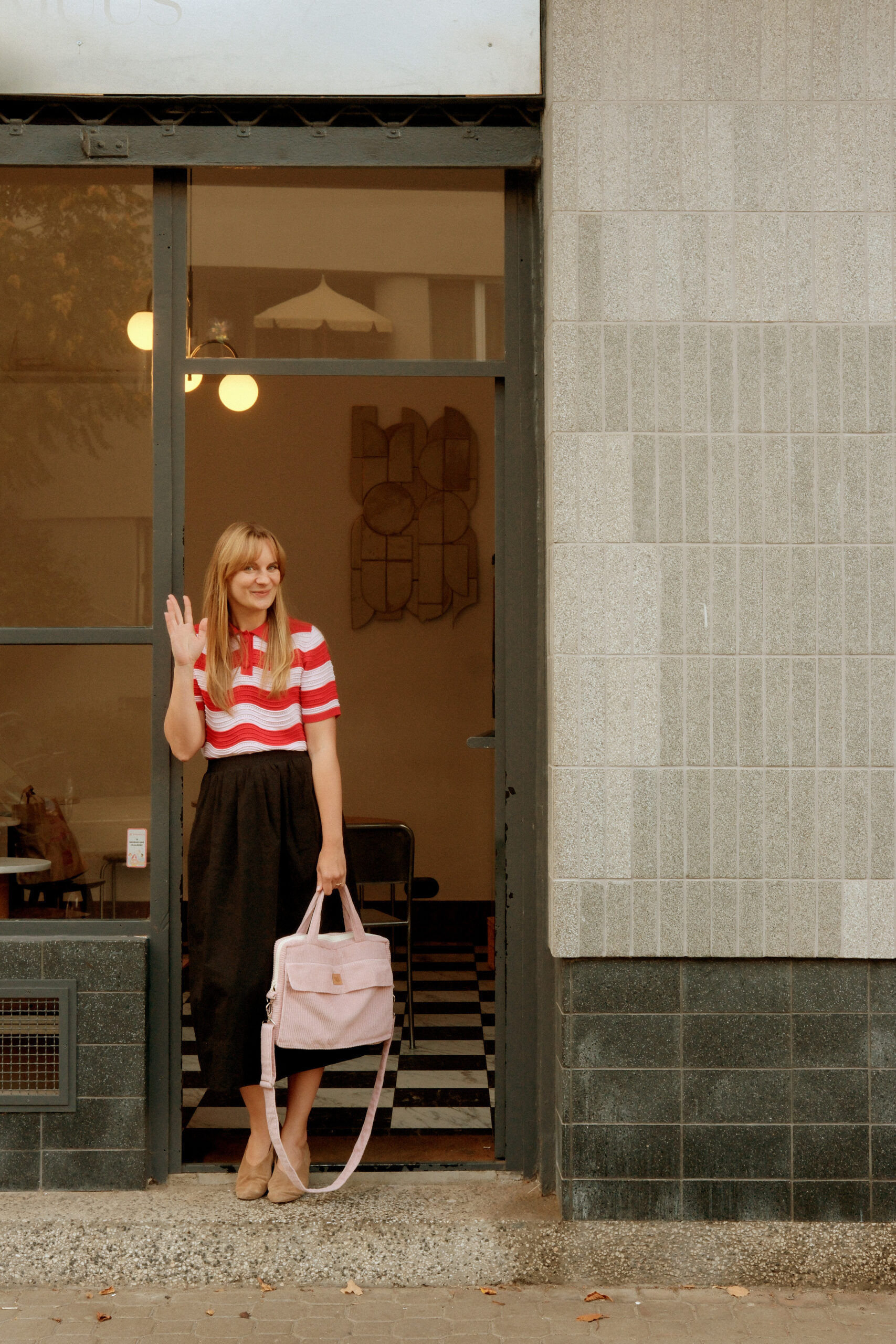 Kobieta stojąca w otwartych drzwiach budynku, uśmiechająca się i machająca ręką, ubrana w czerwono-białą koszulkę w paski i czarną spódnicę, trzymająca różową torbę.