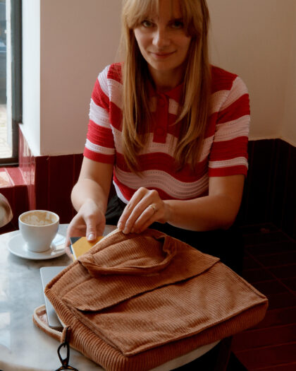 Kobieta w czerwono-białej paskowanej bluzce siedzi przy okrągłym białym stole w kawiarni i wkłada laptop do brązowej, welurowej torby na laptopa, na stole stoi filiżanka po kawie.