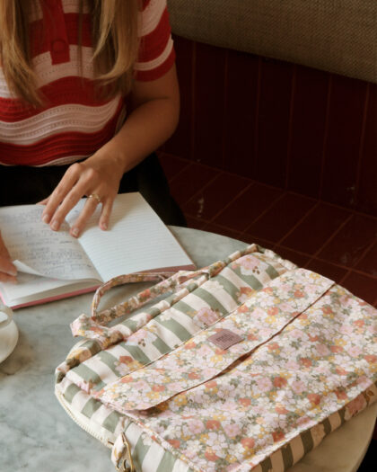 Osoba w czerwono-białej koszulce w paski siedzi przy okrągłym marmurowym stole, przekładając kartki w notatniku, obok leży kwiatowa torba z zielono-białymi pasami i kwiecistym wzorem.
