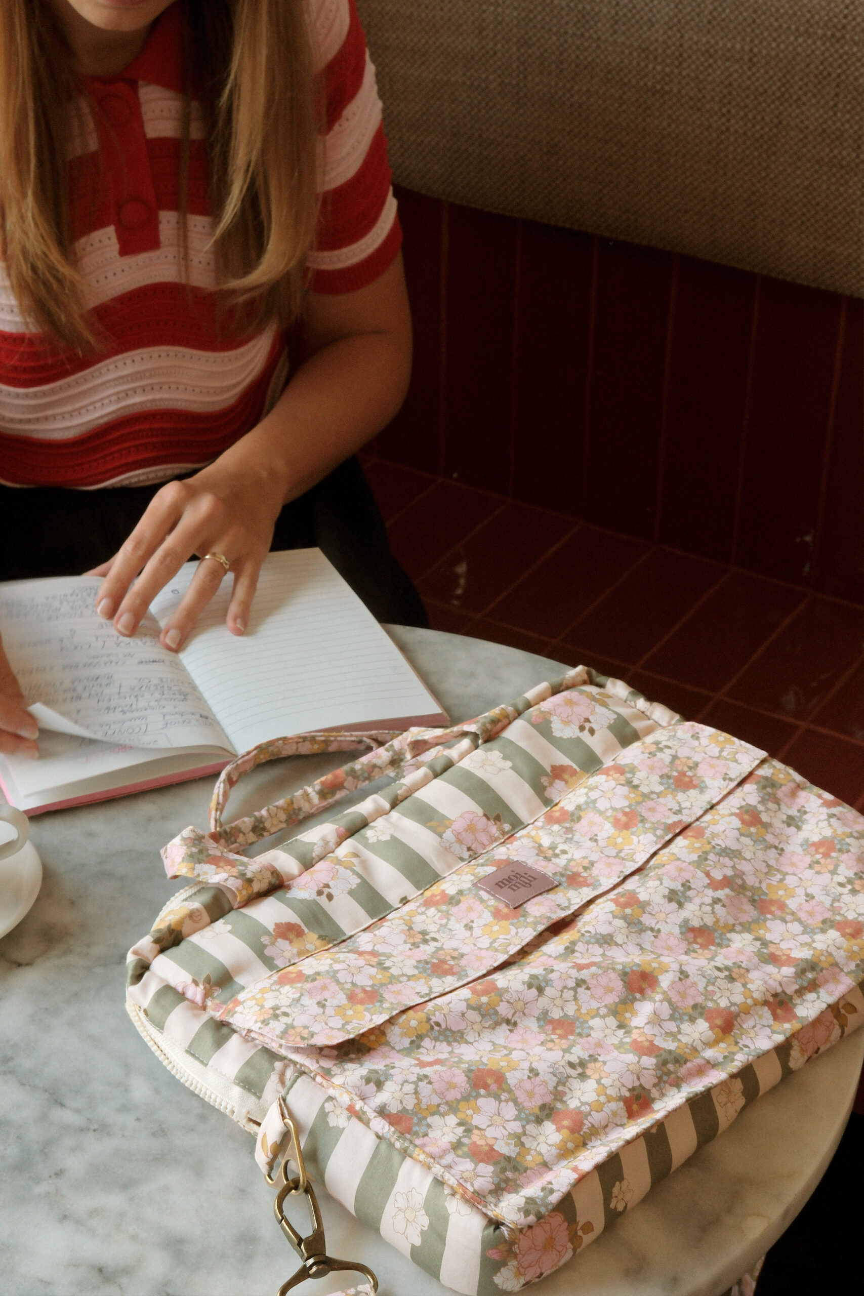 Osoba w czerwono-białej koszulce w paski siedzi przy okrągłym marmurowym stole, przekładając kartki w notatniku, obok leży kwiatowa torba z zielono-białymi pasami i kwiecistym wzorem.