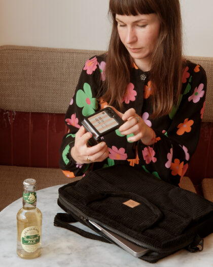 Kobieta w czarnej bluzce z kolorowymi kwiatowymi wzorami siedzi przy okrągłym marmurowym stole, trzymając przenośne urządzenie z wyświetlaczem w dłoniach, na stole stoi butelka napoju Ginger Ale, a obok leży czarna pikowana torba z częściowo wysuniętym laptopem.