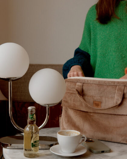 Kobieta w zielonym swetrze stoi przy okrągłym marmurowym stole z lampą, butelką ginger ale, filiżanką kawy i torebką z miękkiego, beżowego materiału.