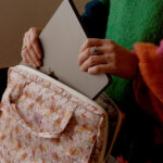 Osoba w kolorowym swetrze wkłada srebrnego laptopa do kwiatowej torby z suwakiem i uchwytem.