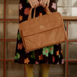 Dłoń trzymająca dużą, brązową torbę z materiału sztruksowego, ubrana w sukienkę w kolorowe kwiaty na czarnym tle, stojąca na czarno-białej podłodze w kratę.