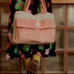 Kobieta w sukience w kolorowe kwiaty trzyma za uchwyty różowo-beżową torbę z miękkiego materiału na tle szklanych bloków i szachownicowej podłogi.