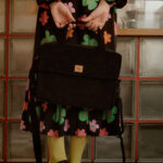 Osoba w sukience z dużymi kolorowymi kwiatami trzyma z tyłu czarną torbę na ramię, stoi na czarno-białej polowej podłodze w kuchni lub kawiarni z szafkami z matowymi szybami w tle.