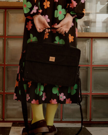 Osoba w sukience z dużymi kolorowymi kwiatami trzyma z tyłu czarną torbę na ramię, stoi na czarno-białej polowej podłodze w kuchni lub kawiarni z szafkami z matowymi szybami w tle.