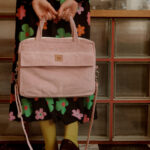 Kobieta stojąca na czarno-białej szachownicy podłodze trzyma różową, prążkowaną torbę z materiału sztruksowego, ubrana w czarną sukienkę w kolorowe kwiaty i żółte skarpetki z czarnymi butami.