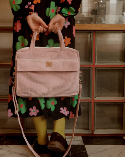 Kobieta stojąca na czarno-białej szachownicy podłodze trzyma różową, prążkowaną torbę z materiału sztruksowego, ubrana w czarną sukienkę w kolorowe kwiaty i żółte skarpetki z czarnymi butami.