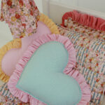 Dwa sercowate poduszki w kratkę z falbanką w kolorze różowym i żółtym na kwiecistym i pasiastym kołdrze na białym łóżku, obok zwisający kawałek tkaniny w kwiatowy wzór.