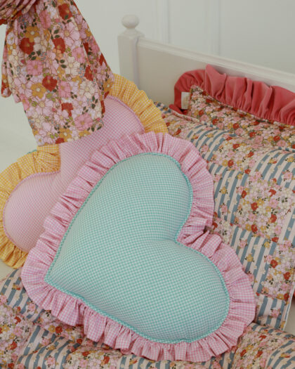 Dwa sercowate poduszki w kratkę z falbanką w kolorze różowym i żółtym na kwiecistym i pasiastym kołdrze na białym łóżku, obok zwisający kawałek tkaniny w kwiatowy wzór.