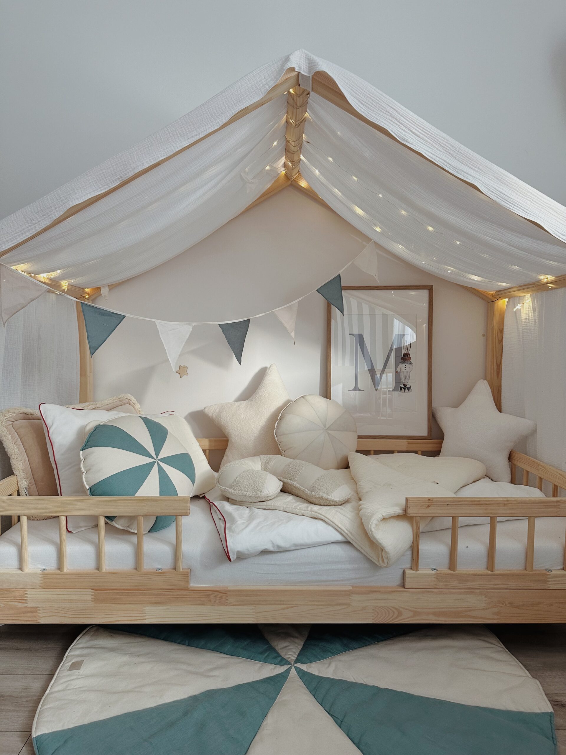 Drewniane łóżko dziecięce z baldachimem z białej tkaniny ozdobione lampkami i girlandą z materiałowych trójkątnych chorągiewek, na łóżku poduszki w kształcie gwiazdy i koła oraz jasna pościel, a pod łóżkiem okrągły dywan w biało-zielonego wzoru.