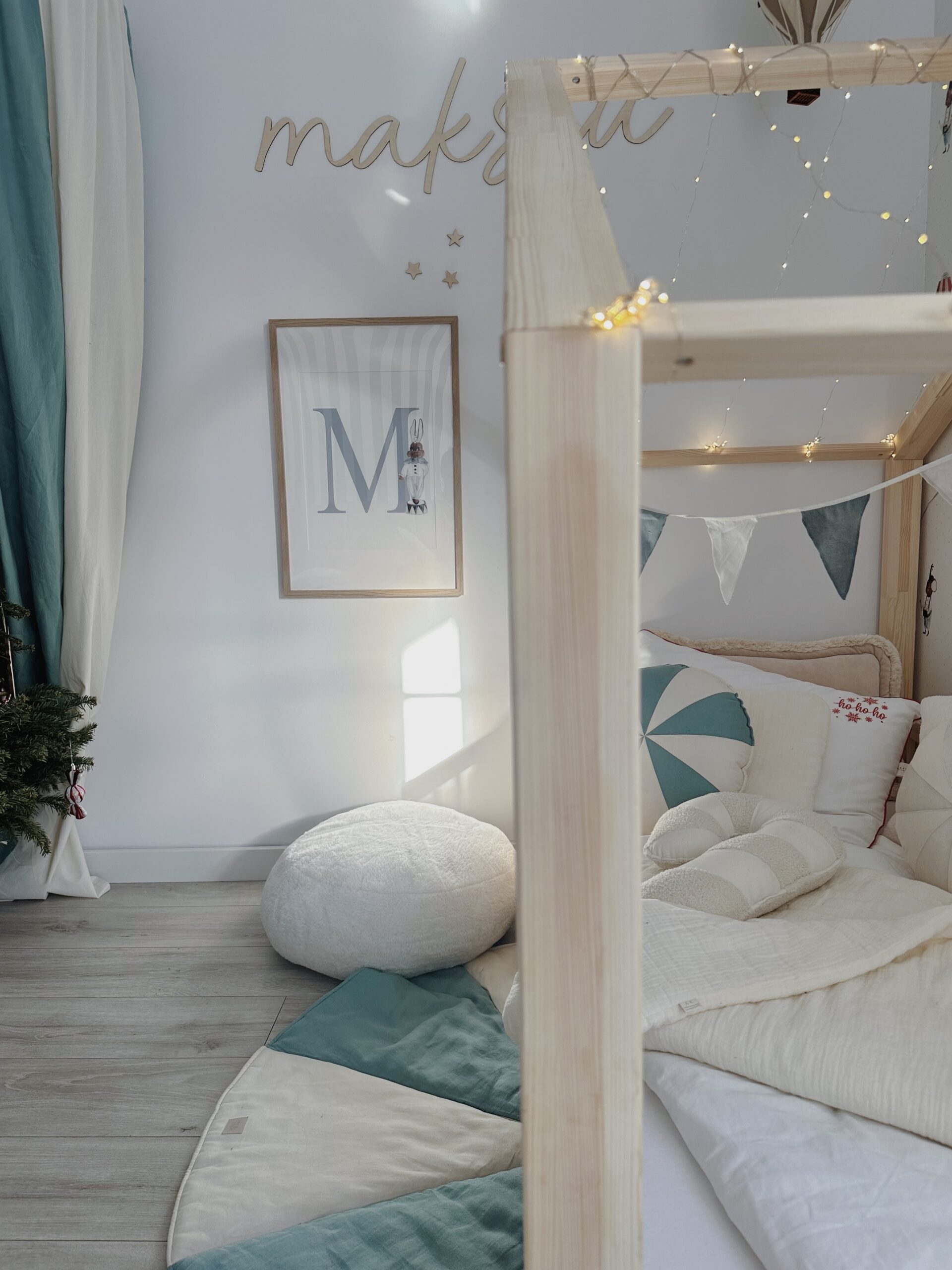 Przytulny pokój dziecięcy z jasnym, drewnianym łóżkiem w kształcie domku ozdobionym lampkami, podłoga wyłożona dywanem i białą pufą, na ścianie obraz z literą M i zawieszonym króliczkiem, część pomieszczenia udekorowana zasłonami w odcieniach niebieskiego i białego.