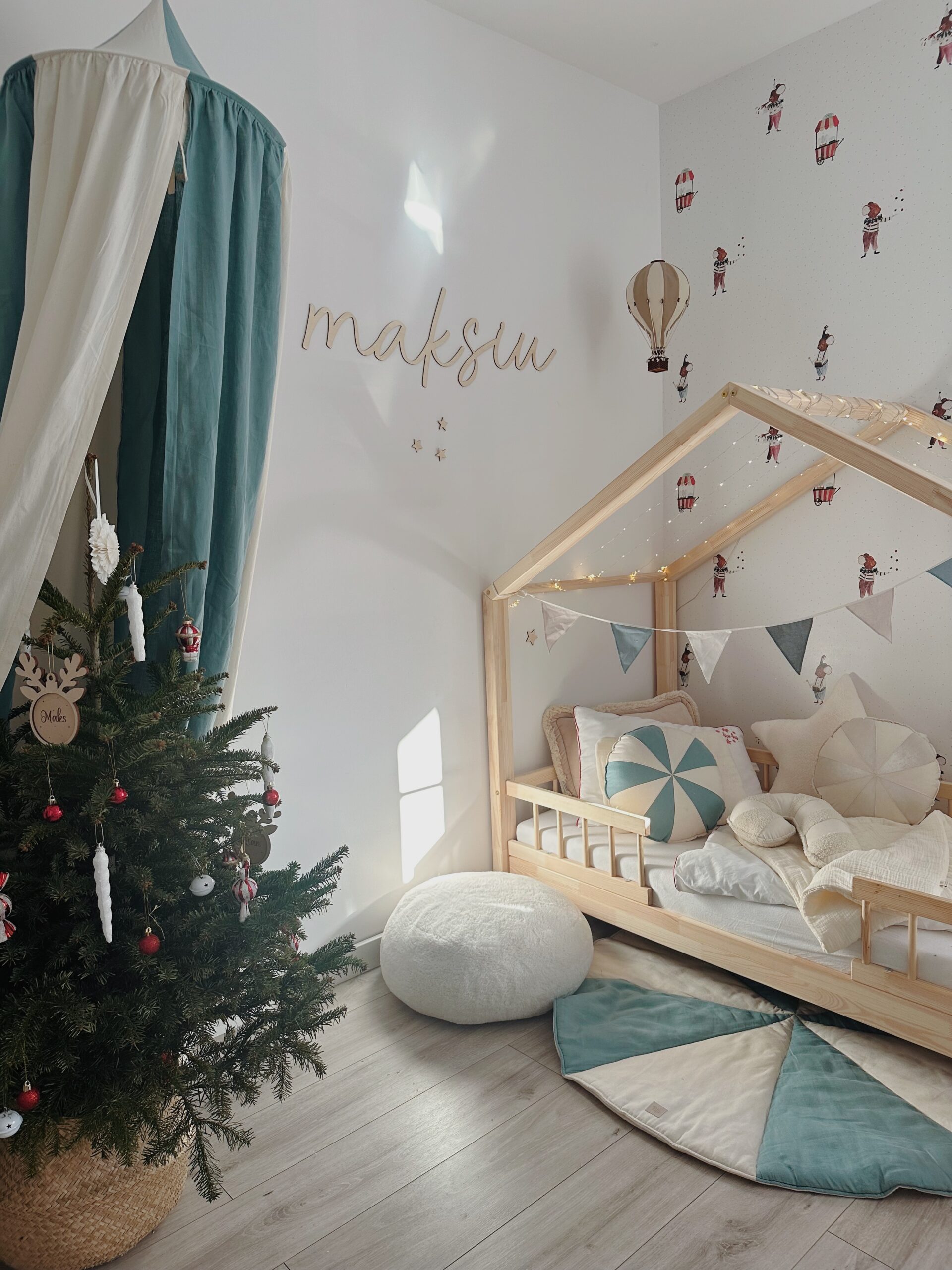 Pokój dziecięcy z drewnianym łóżkiem w kształcie domku, ozdobionym girlandą i poduszkami, obok dekoracyjny namiot w kolorach kremowo-zielonych, choinka z czerwonymi i białymi ozdobami, na ścianie napis maksu i tapeta z motywem postaci i balonów powietrznych.