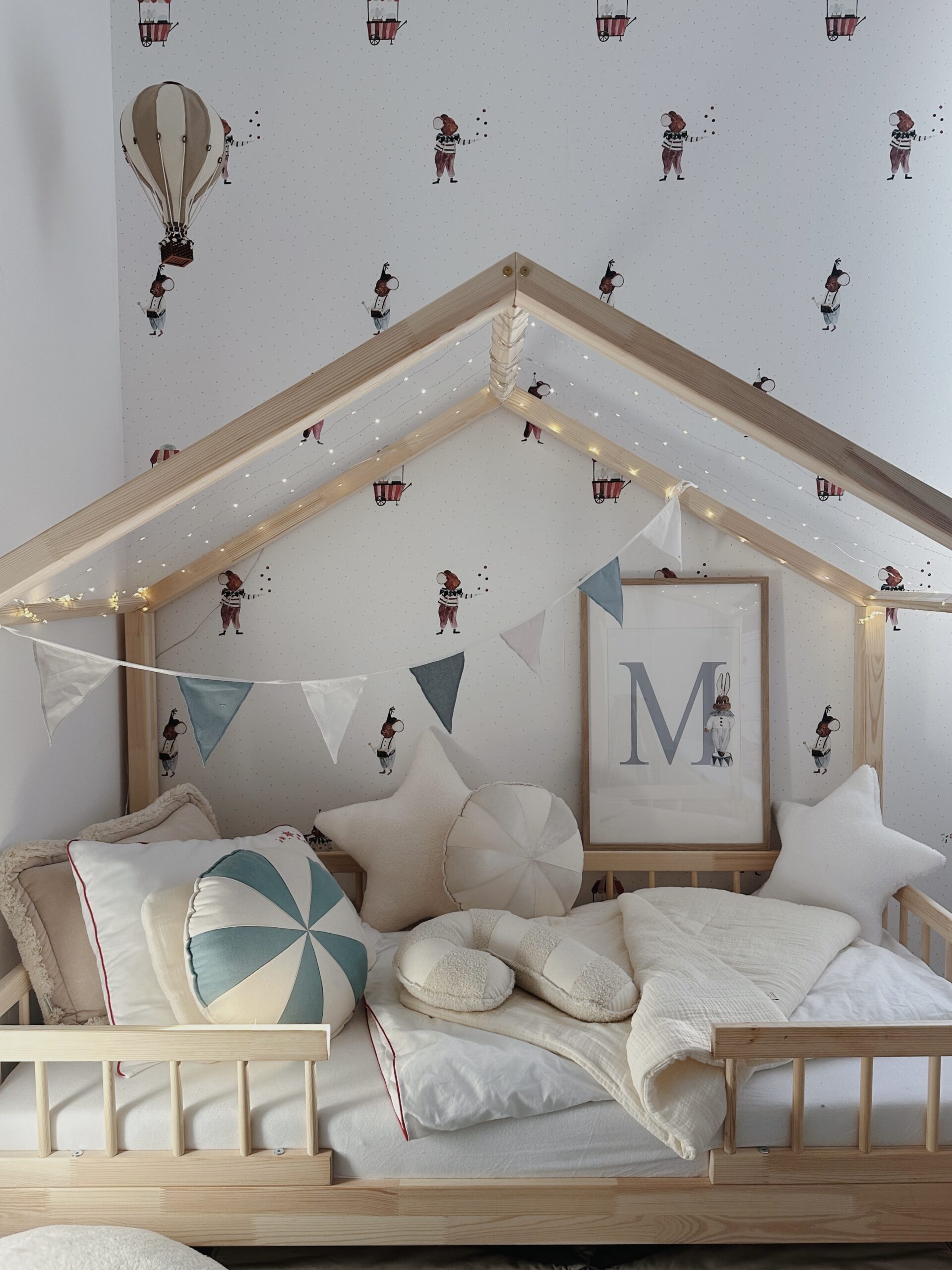 Łóżko dziecięce w kształcie domku z drewnianą ramą i girlandą z lampkami, ozdobione poduszkami w kształcie gwiazd, kółka i croissant, na tle tapety z ilustracjami postaci i balonów.