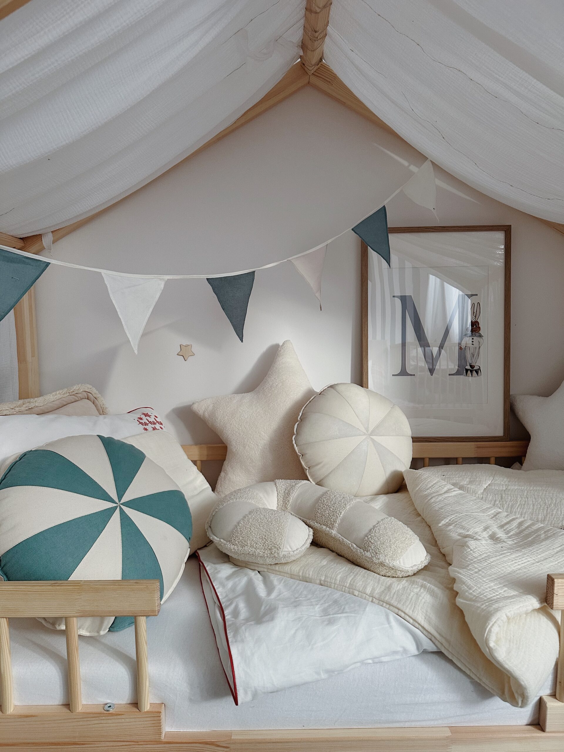 Pokój dziecięcy z łóżkiem w drewnianej ramie i białym baldachimem, udekorowany poduszkami w kształcie gwiazdy i okrągłymi, flagami w odcieniach bieli i zieleni zawieszonymi na ścianie oraz dużym obrazem z literą M w drewnianej ramie.