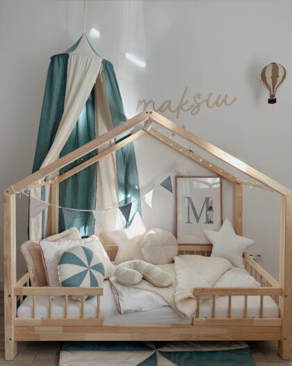 Drewniane łóżko dziecięce w kształcie domku udekorowane światełkami i girlandą z trójkątnych flag w odcieniach bieli, szarości i zieleni, z poduszkami w kształcie gwiazdek i okrągłej oraz narzutą w jasnych kolorach, za łóżkiem zielono-biała baldachimowa zasłona i ściana ozdobiona drewnianym napisem "maksiu", obrazem z literą M i figurką królika oraz wiszącym balonem dekoracyjnym, wszystko w jasnym, minimalistycznym pokoju dziecięcym.
