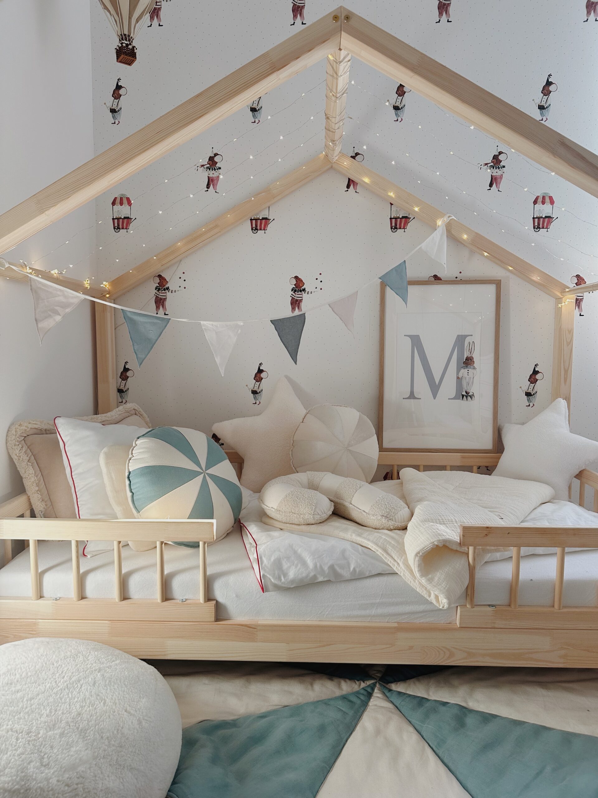 Drewniane łóżko w kształcie domku z jasną pościelą i dekoracyjnymi poduszkami w kształcie gwiazdy, okrągłej i księżyca, zawieszone girlandy z trójkątnych flag w pastelowych kolorach, tapeta z wzorem postaci zwierząt i balonów oraz plakat z literą M w ramce na ścianie.