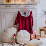 Czerwone dziecięce sukienka z białą koronkową kryzą na wieszaku na tle białych mebli, otoczona białymi ozdobnymi poduszkami, miękkim dywanem, pluszową myszką w czerwonym czapce, prezentem i pomarańczowymi światełkami LED.