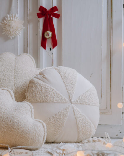 Trzy miękkie poduszki o kształtach chmur i okrągłej, geometrycznej formy na jasnym dywanie przed starymi, białymi drzwiami ozdobionymi czerwonym kokardkiem i ozdobą z papieru, z delikatnym oświetleniem girlandy świetlnej rozłożonej na podłodze