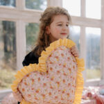 Dziewczynka z jasnobrązowymi kręconymi włosami trzymająca duża poduszkę w kształcie serca z kwiatowym wzorem i żółtą falbanką, stojąca w jasnym pomieszczeniu z dużymi oknami i girlandą żółtych proporczyków w tle.