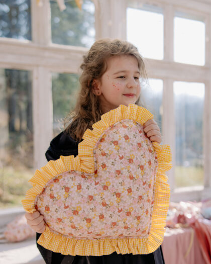 Dziewczynka z jasnobrązowymi kręconymi włosami trzymająca duża poduszkę w kształcie serca z kwiatowym wzorem i żółtą falbanką, stojąca w jasnym pomieszczeniu z dużymi oknami i girlandą żółtych proporczyków w tle.