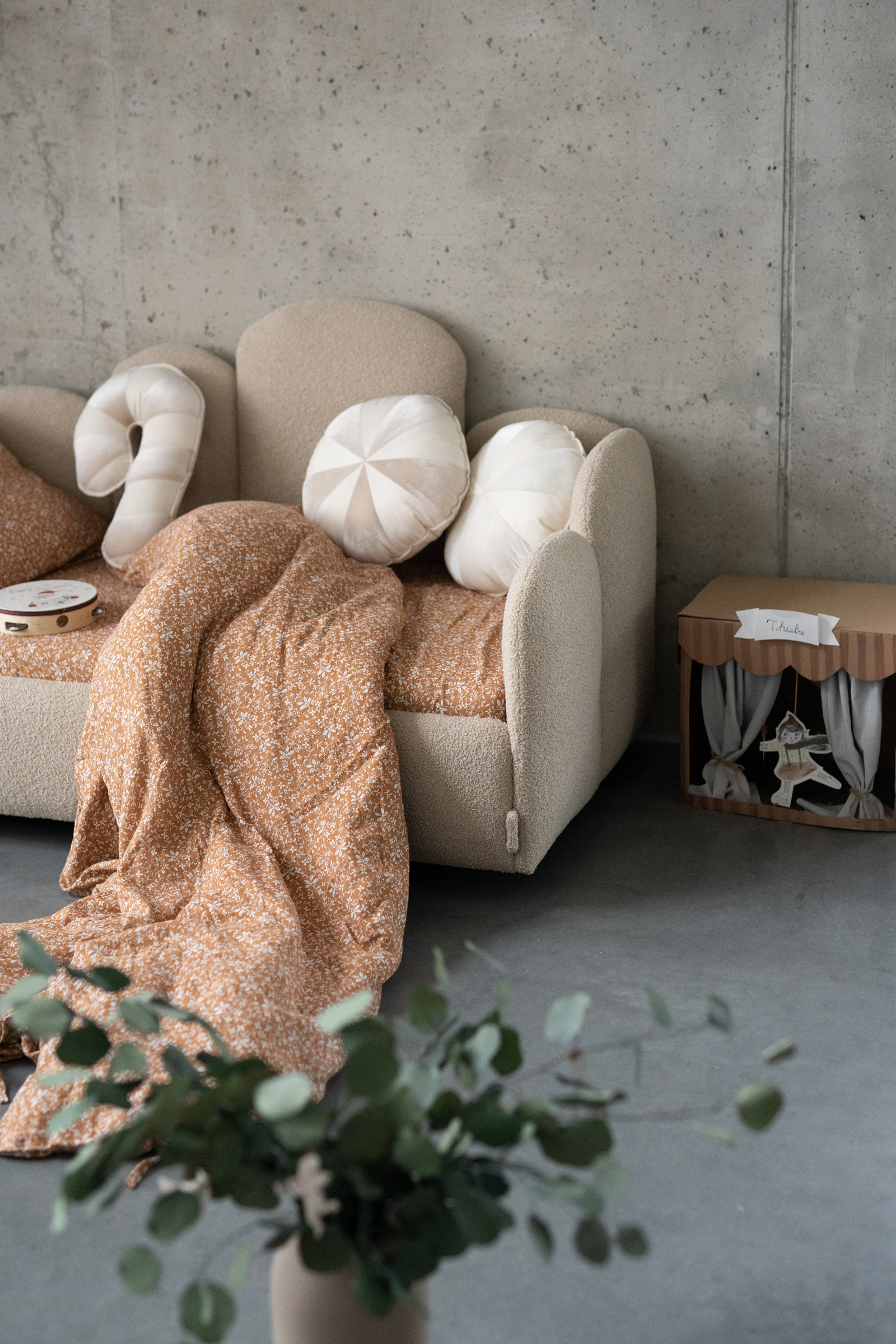 Beżowa sofa dziecięca z beżowo-białymi poduszkami w kształcie lizaków, pomarańczowy koc w drobne białe wzory rozwieszony na sofie, obok kartonowy teatrzyk dla dzieci na tle szarej ściany betonowej, w pierwszym planie rozmyte liście rośliny w doniczce.