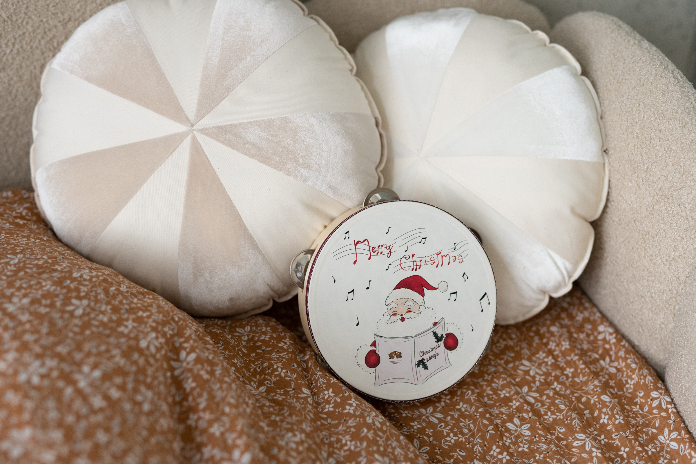 Okrągła drewniana tamburyna z obrazkiem Świętego Mikołaja i napisem Merry Christmas, ustawiona na pomarańczowej narzucie z białym wzorem, między dwoma beżowo-białymi, okrągłymi poduszkami na kanapie.