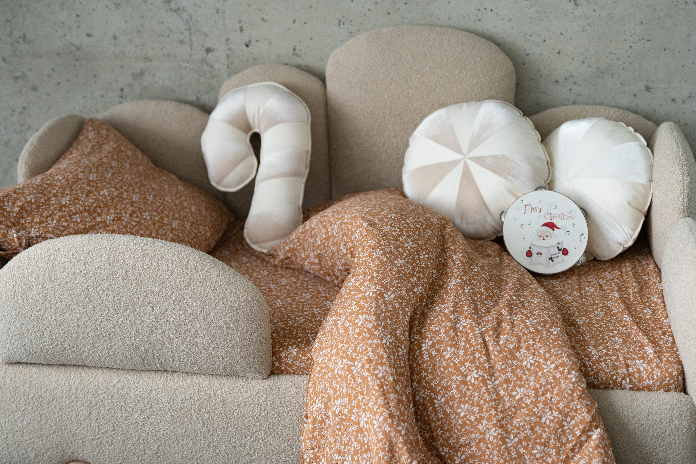 Beżowa teksturowana sofa z brązową narzutą w białe kwiatowe wzory oraz kilkoma poduszkami, w tym okrągłymi kremowymi i jedną w kształcie laski cukrowej, na jednej z poduszek ozdoba świąteczna z Mikołajem i napisem Merry Christmas