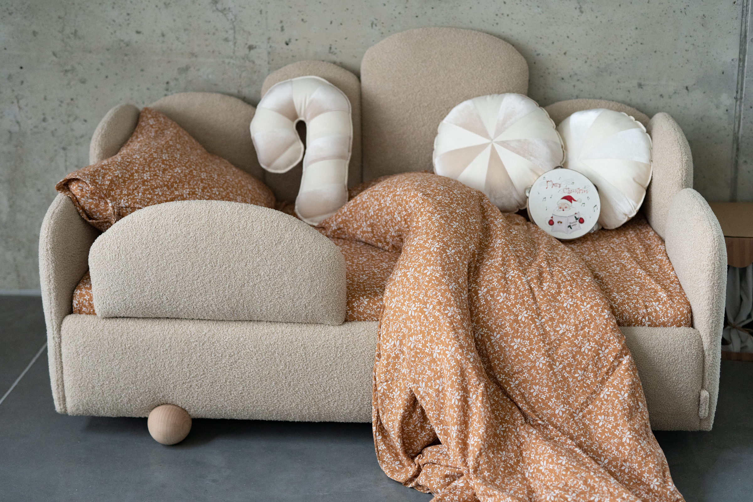 Mała beżowa kanapa dziecięca z teksturowaną tkaniną, udekorowana poduszkami w kształcie lizaka i okrągłymi poduszkami w jasnych odcieniach, z pomarańczową narzutą w białe kwiaty, stojąca na szarym podłożu przed betonową ścianą.