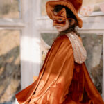 Dziewczynka w pomarańczowym kostiumie czarownicy z kapeluszem, maską na oczy i peleryną z cekinowym kapturem, trzymająca biały gwiazdkowy różdżkę, stoi bokiem i spogląda przez ramię w stronę aparatu, w tle jasne okna i pastelowe girlandy z trójkątnymi flagami.