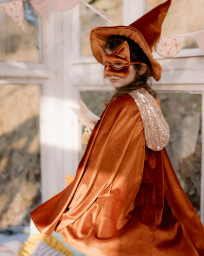 Dziewczynka w pomarańczowym kostiumie czarownicy z kapeluszem, maską na oczy i peleryną z cekinowym kapturem, trzymająca biały gwiazdkowy różdżkę, stoi bokiem i spogląda przez ramię w stronę aparatu, w tle jasne okna i pastelowe girlandy z trójkątnymi flagami.