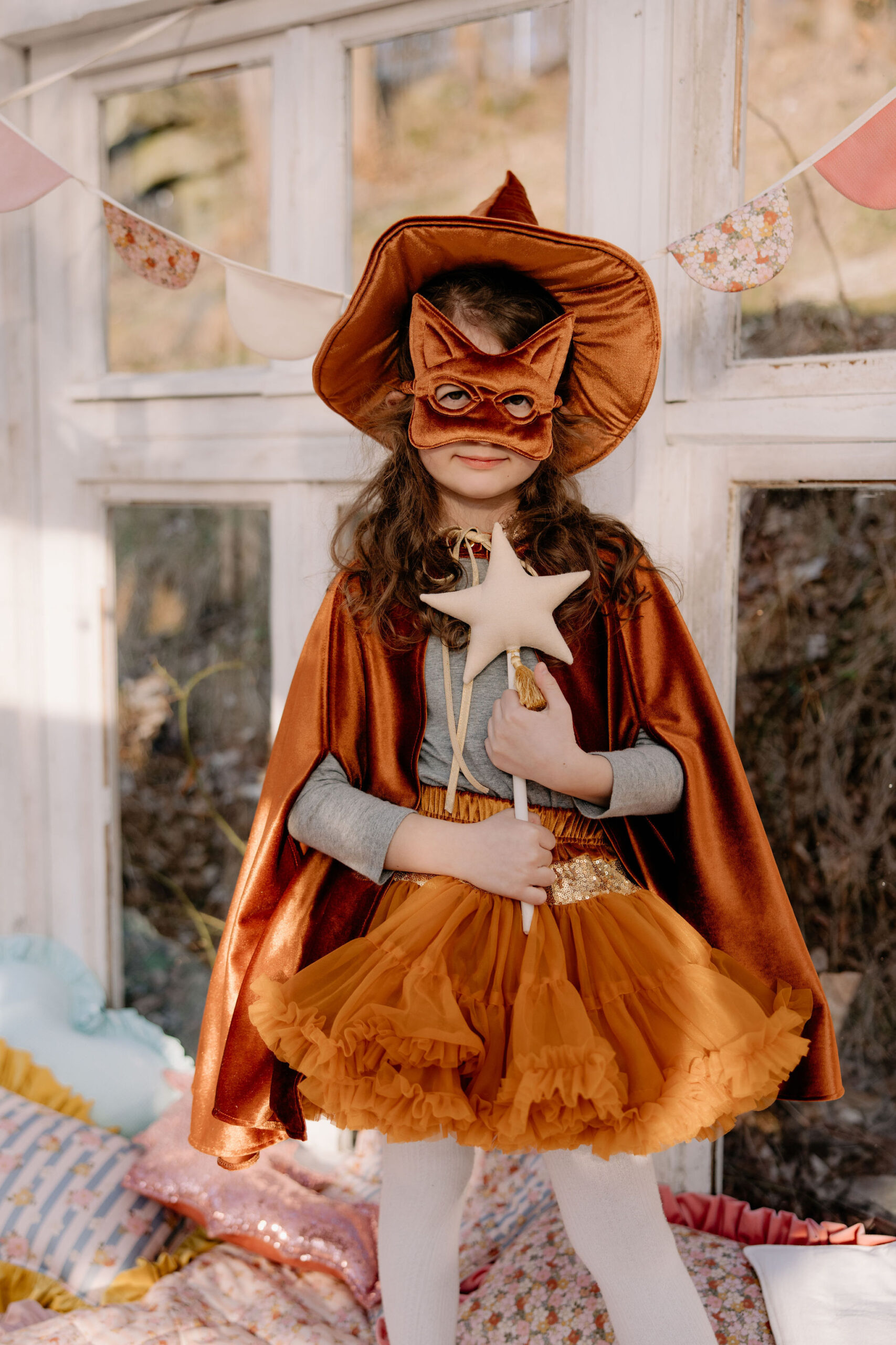 Dziewczynka w pomarańczowym kostiumie czarownicy z peleryną, kapeluszem i maską w kształcie kota, trzymająca gwiezdną różdżkę, stojąca w jasnym pokoju z dekoracjami i poduszkami na podłodze.