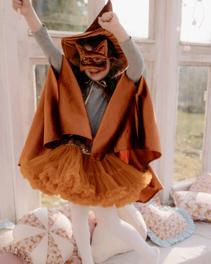Dziewczynka w pomarańczowym kostiumie kota z peleryną, maską i tiulową spódniczką, stojąca na łóżku z poduszkami, unosząca ręce w górę w jasnym pomieszczeniu z dużymi oknami.
