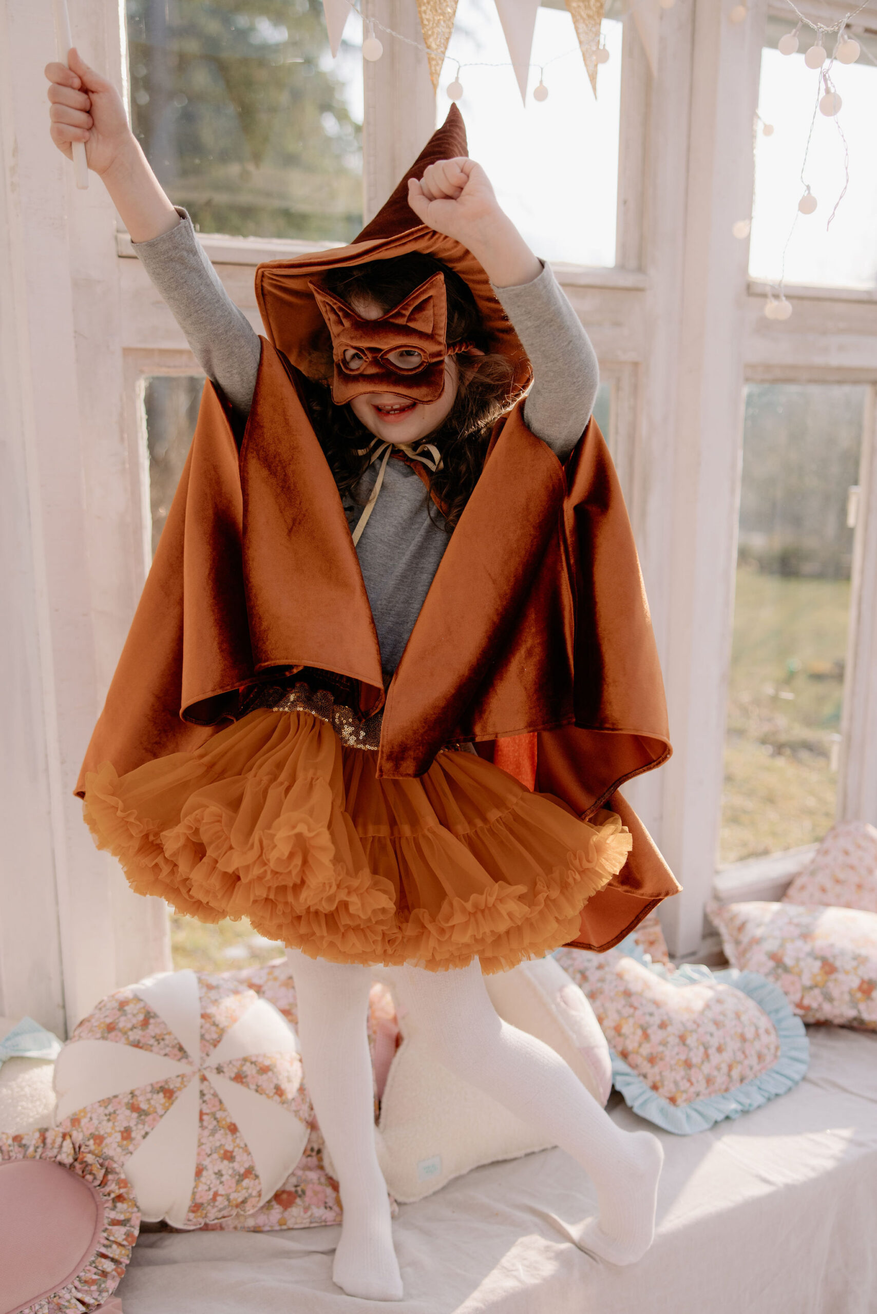 Dziewczynka w pomarańczowym kostiumie kota z peleryną, maską i tiulową spódniczką, stojąca na łóżku z poduszkami, unosząca ręce w górę w jasnym pomieszczeniu z dużymi oknami.