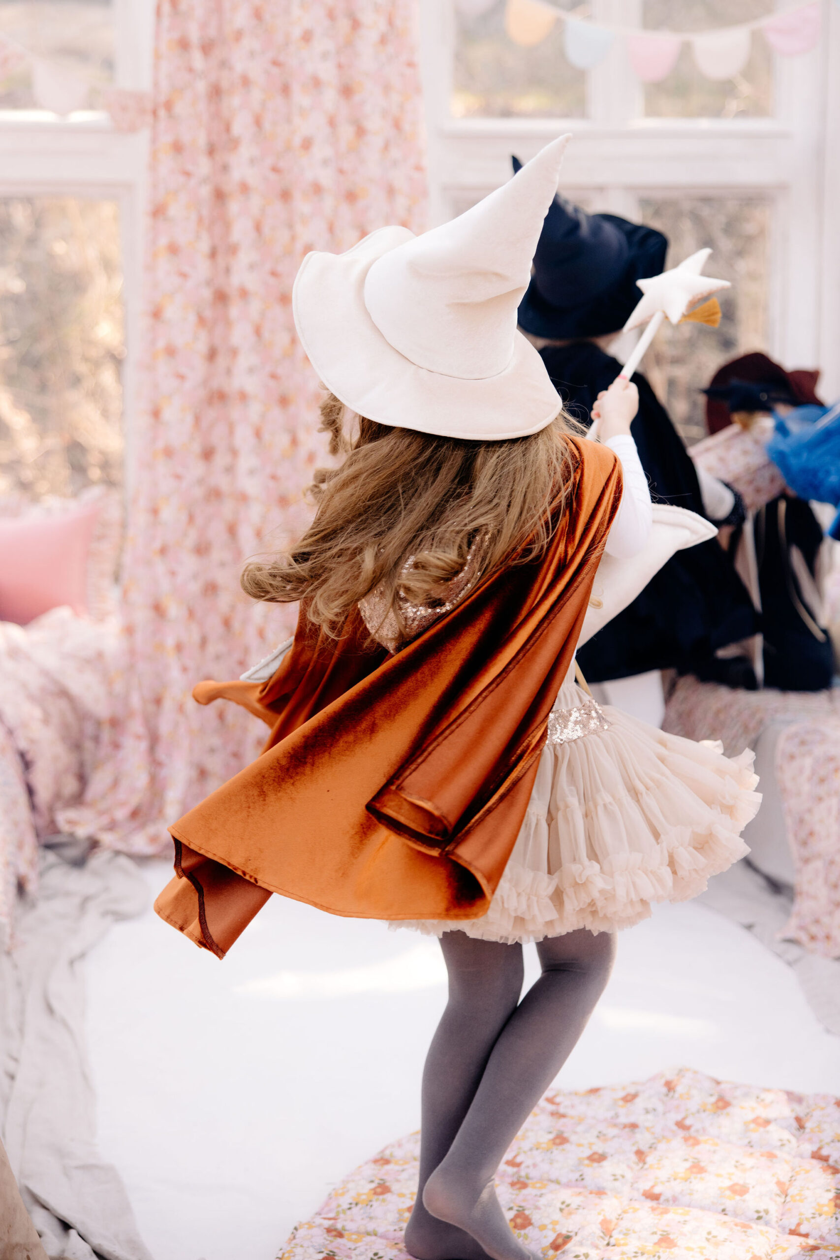 Dziewczynka w białym kapeluszu czarownicy i pomarańczowej pelerynie, trzymająca różdżkę z gwiazdą, stojąca na tle jasnego pokoju z kwiecistymi zasłonami i innymi dziećmi w strojach czarodziei.