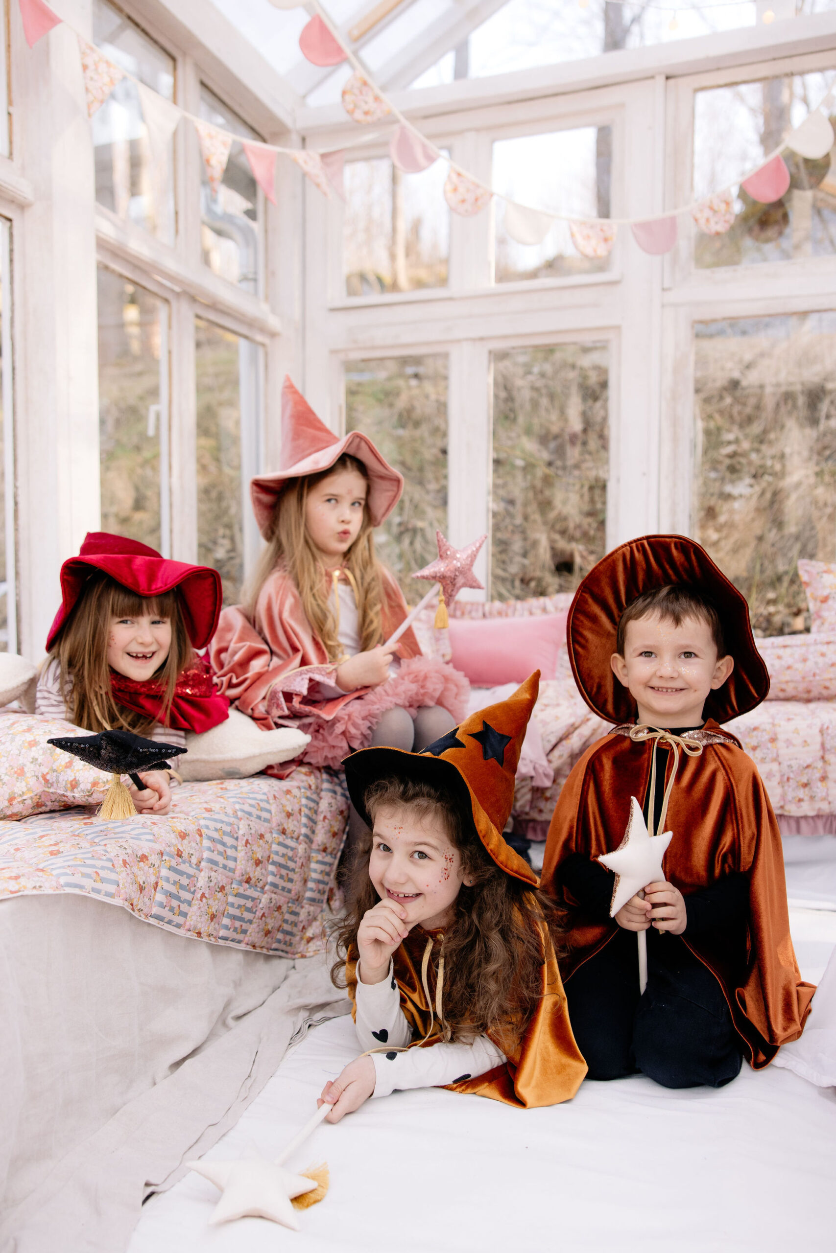 Cztery dzieci w kolorowych kostiumach czarodziei i czarownic bawiące się w jasnym pokoju z dużymi oknami, dwie dziewczynki siedzą na kanapie, a chłopiec i dziewczynka leżą na podłodze, trzymając różdżki w kształcie gwiazdek.