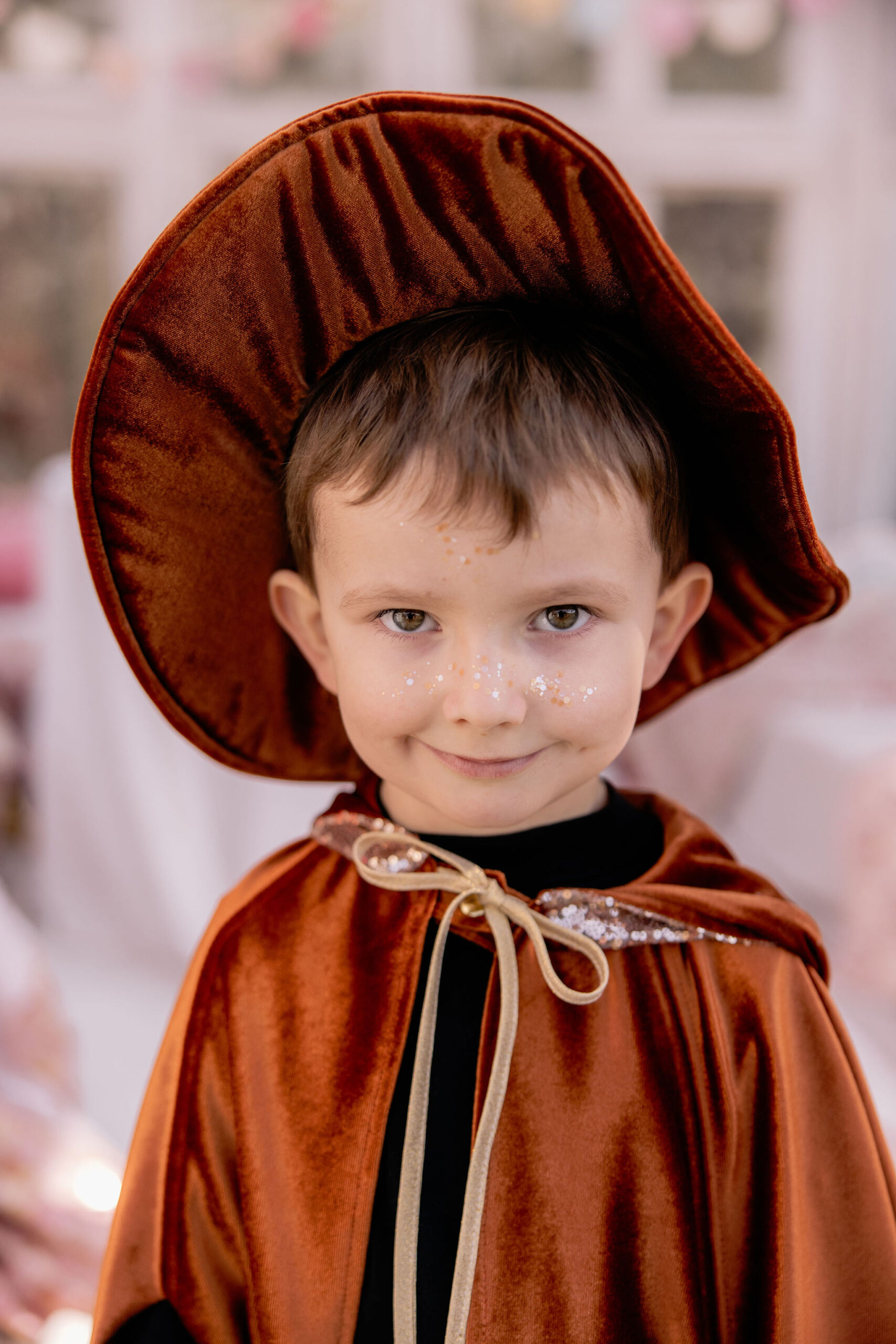 Portret uśmiechniętego chłopca w kostiumie czarodzieja z rudą aksamitną peleryną i dużym kapeluszem, z błyszczącymi ozdobami na twarzy.