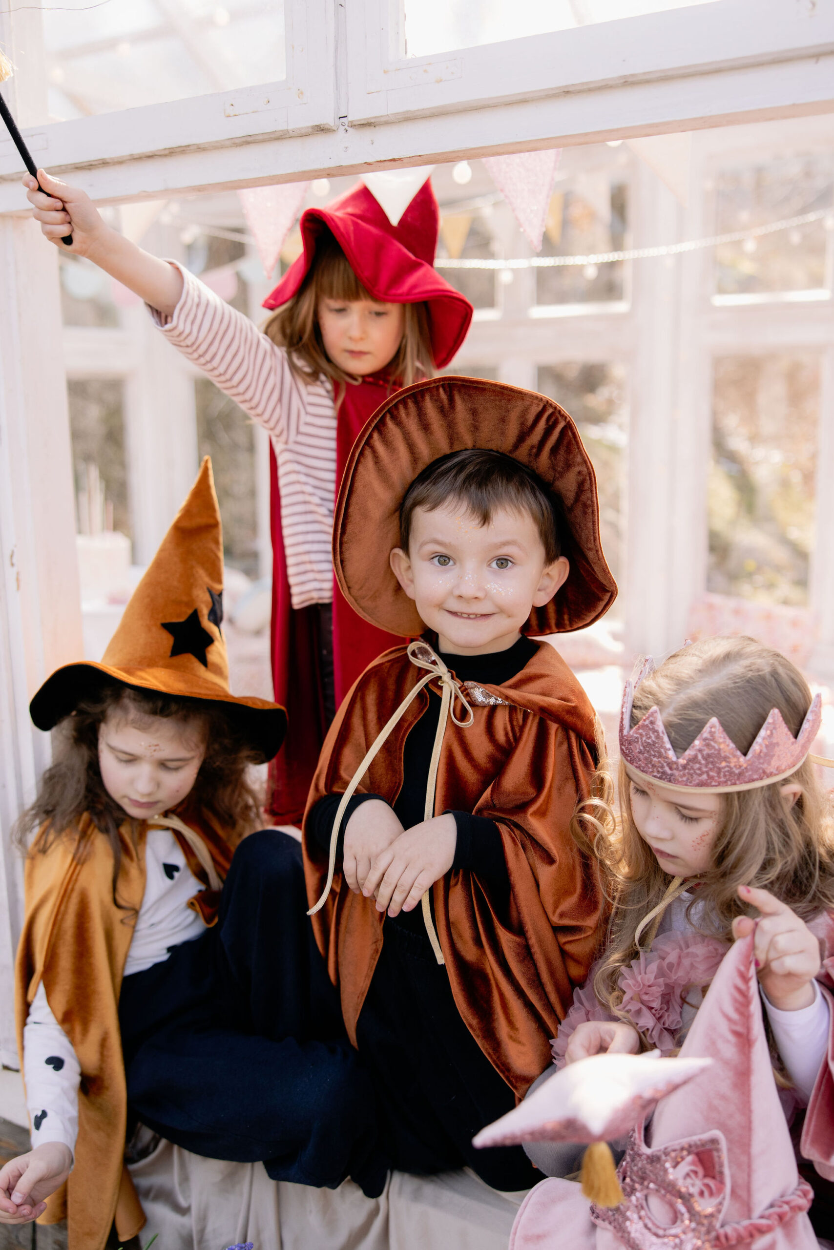 Cztery dzieci w kolorowych kostiumach bawiące się razem w jasnym pokoju ozdobionym girlandami, jedno z dzieci w czerwonym kapeluszu trzyma różdżkę, drugie w pomarańczowym kapeluszu czarownicy, trzecie uśmiecha się w brązowym kostiumie, a czwarte nosi różową koronę i zabawkę.