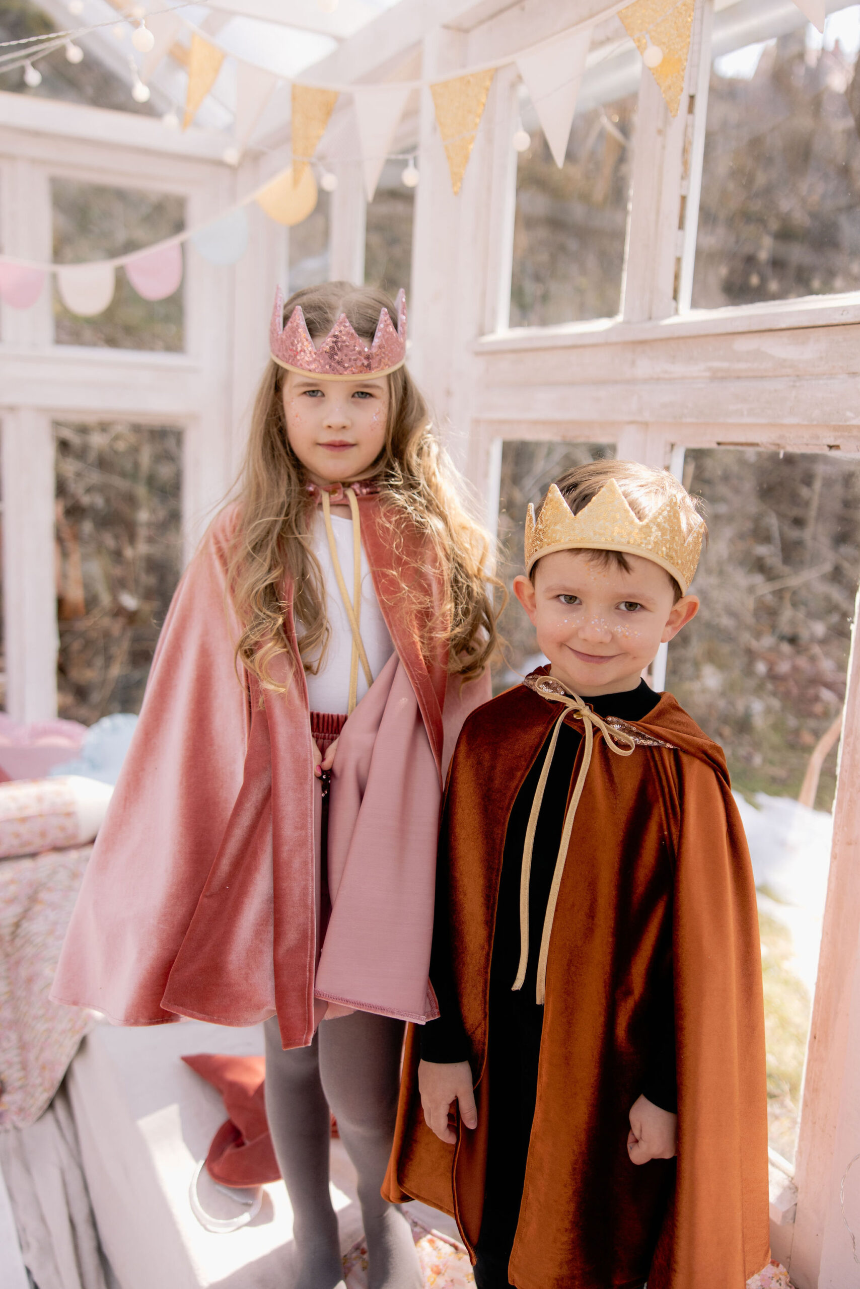 Dwoje dzieci w królewskich kostiumach, dziewczynka w różowym płaszczu i różowej koronie, chłopiec w brązowym płaszczu i złotej koronie, stojący w jasnym, udekorowanym pomieszczeniu z girlandami.