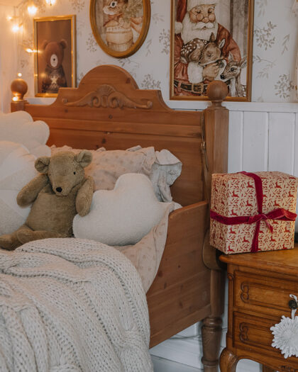 Domek dziecięcy z łóżkiem z drewna ozdobionym pluszowym misiem, miękkimi poduszkami w kształcie serca i kwiatów oraz grubym sweterkowym kocem, obok drewniana szafka nocna z lampką w kształcie motyla, prezentem i świąteczną dekoracją, na ścianie wiszą ilustracje z misiem i Świętym Mikołajem oraz biała sukienka z złotymi gwiazdkami, miękkie ciepłe oświetlenie tworzy przytulną atmosferę.