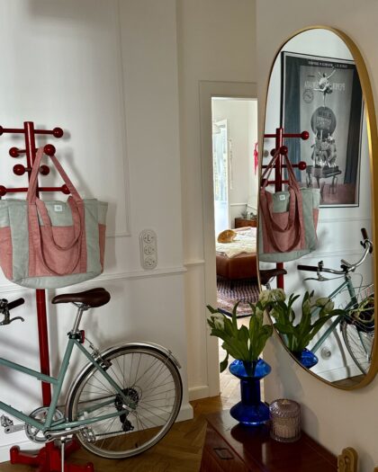 Wnętrze mieszkania z czerwonym stojakiem na rower, zielonym rowerem, torbą na stojaku oraz lustrem odbijającym niebieski wazon z białymi kwiatami.