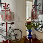 Ścienny czerwony wieszak z różowo-szarym torbą, niebieski wazon z białymi kwiatami na drewnianej konsoli i odzwierciedlenie w lustrze, w tle rower i widok na pokój z pomarańczowym kotem na łóżku.