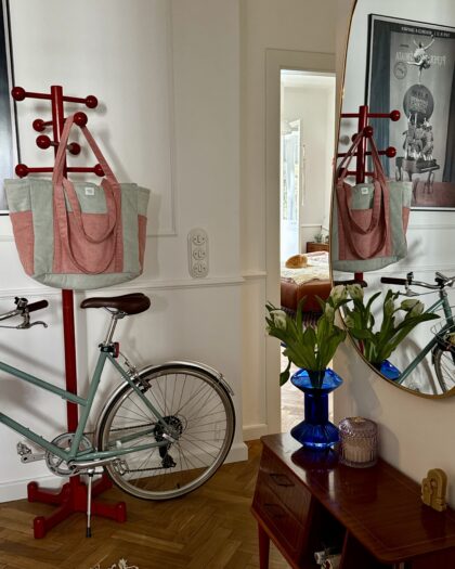 Ścienny czerwony wieszak z różowo-szarym torbą, niebieski wazon z białymi kwiatami na drewnianej konsoli i odzwierciedlenie w lustrze, w tle rower i widok na pokój z pomarańczowym kotem na łóżku.