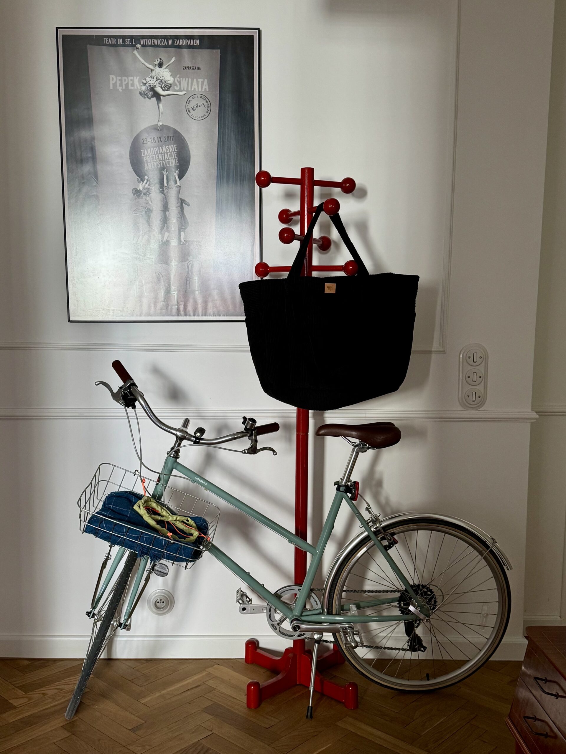 Miętowy rower z koszem na przednim kole, stojący obok czerwonego stojaka na ubrania z czarną torbą, obok biały plakat na ścianie.